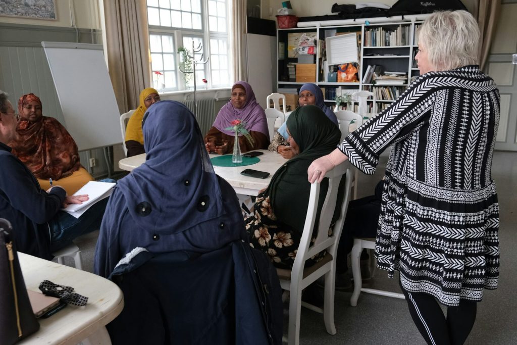 En grupp kvinnor sitter runt ett bord på ett möte medan en kvinna står upp och talar till dem i ett ljust rum.