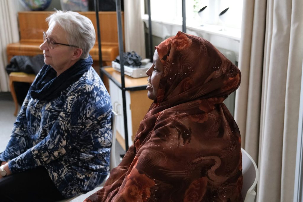 Två kvinnor, den ena i blåmönstrad jacka och den andra i brun hijab, sitter inomhus sida vid sida.