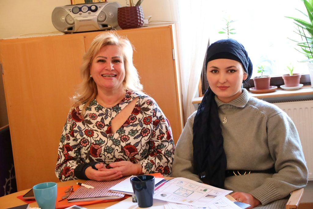 Två kvinnor sitter vid ett bord med papper, muggar och anteckningsböcker och ler i ett mysigt rum med växter.