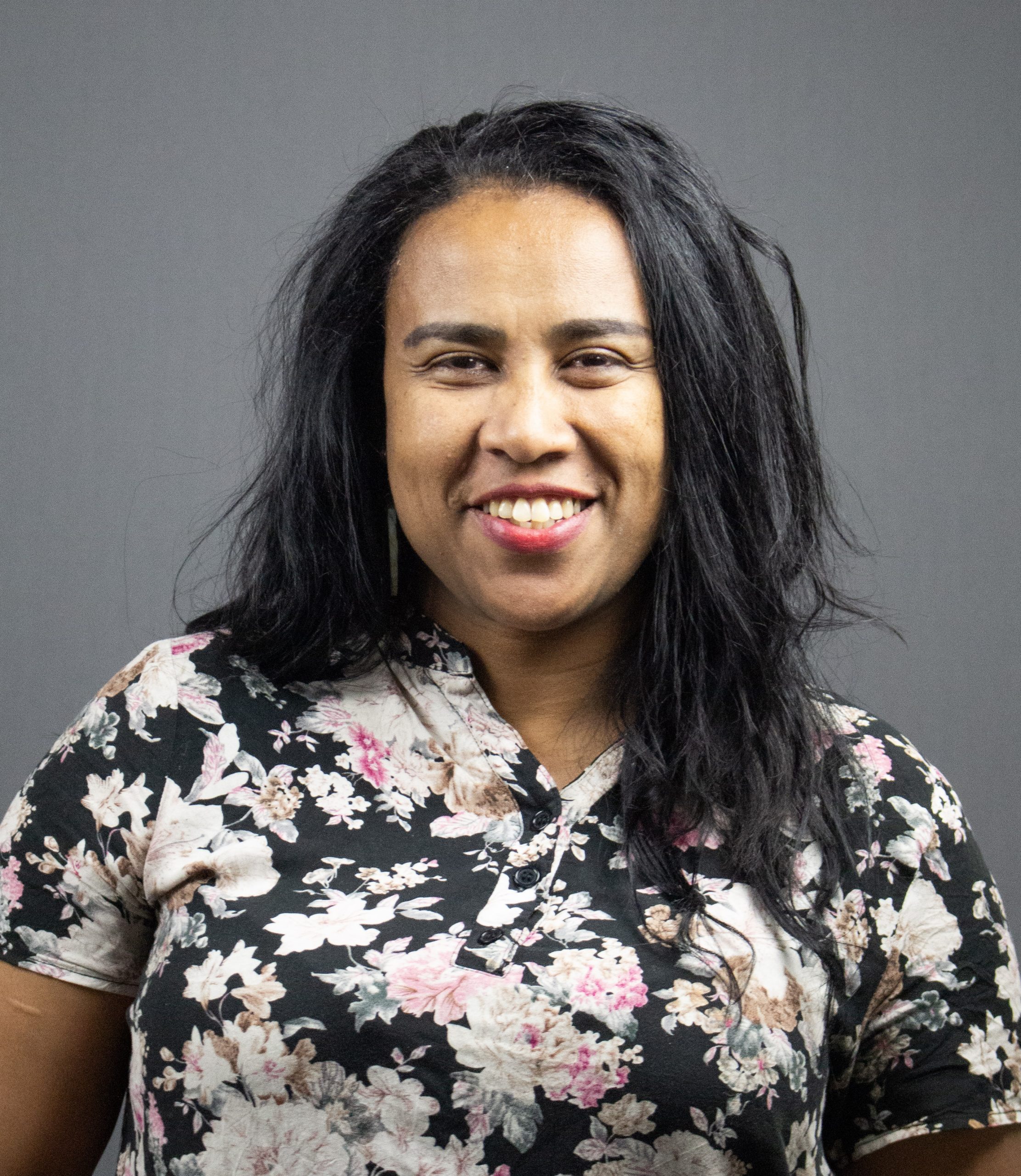 Kvinna med långt mörkt hår, leende, klädd i blommig skjorta, står framför en grå bakgrund.