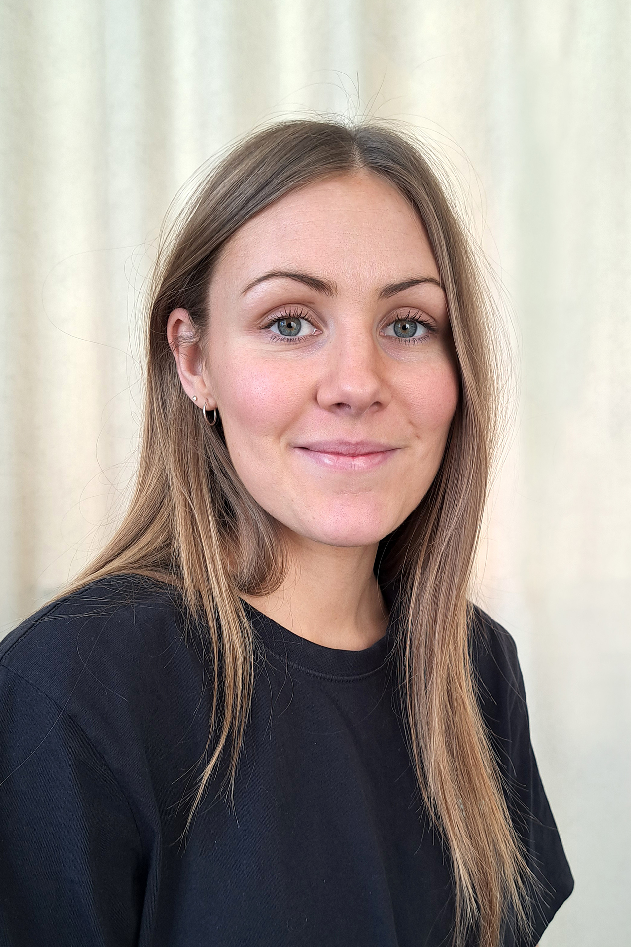 En kvinna med långt ljusbrunt hår och svart skjorta ler mjukt mot en ljus bakgrund.