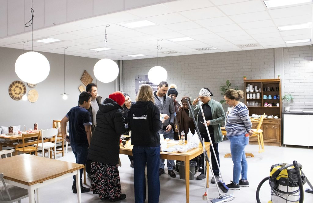 En grupp människor samlas runt ett bord inomhus, några håller i rengöringsmedel och pratar med varandra.