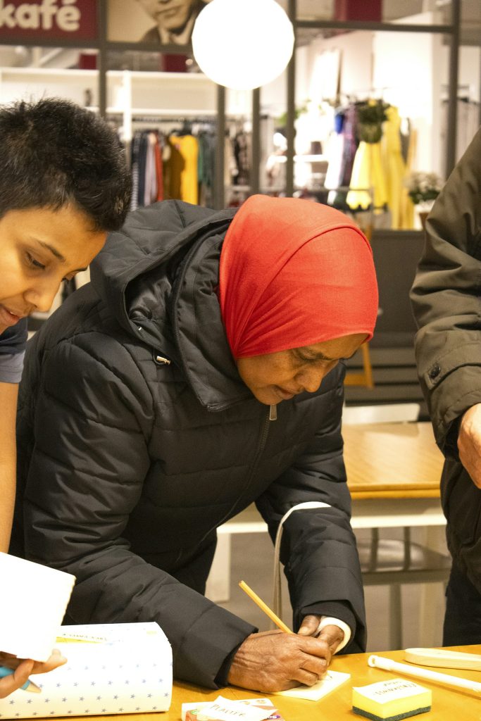 En kvinna med röd huvudduk skriver på ett anteckningsblock vid ett bord inomhus, med människor omkring sig.