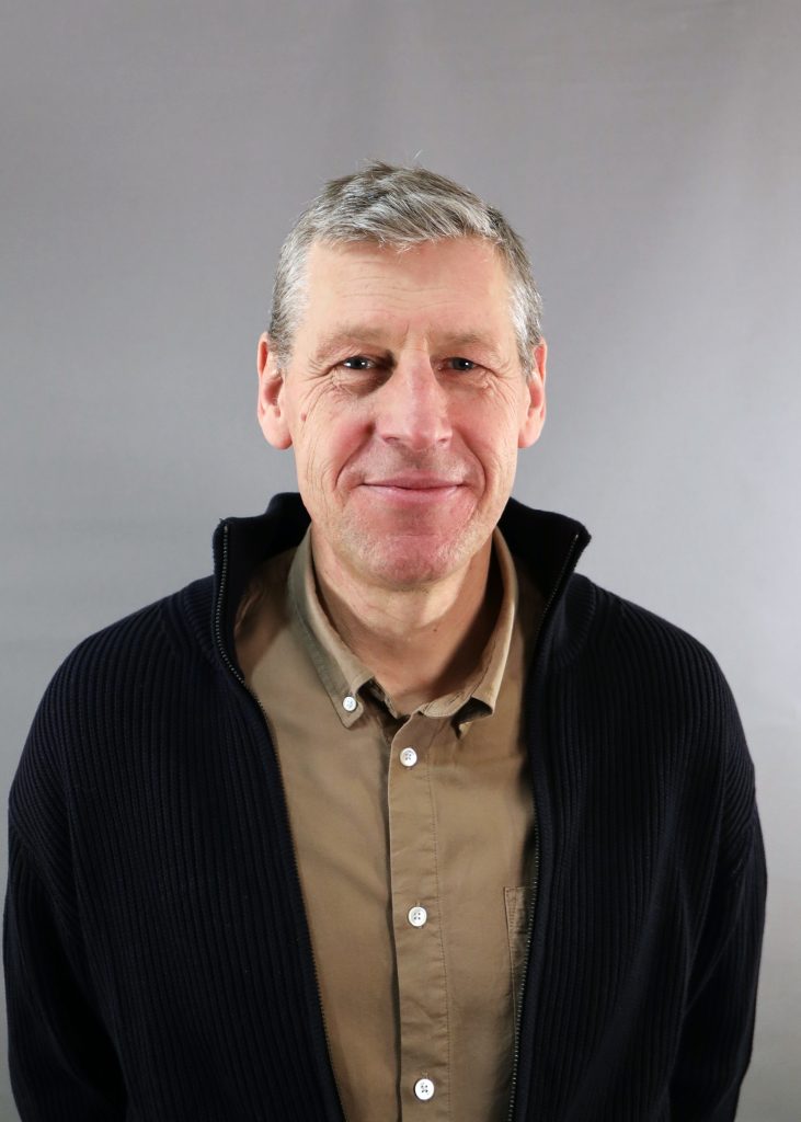 En medelålders man med kort grått hår ler, klädd i brun skjorta och mörk kavaj, mot en grå bakgrund.