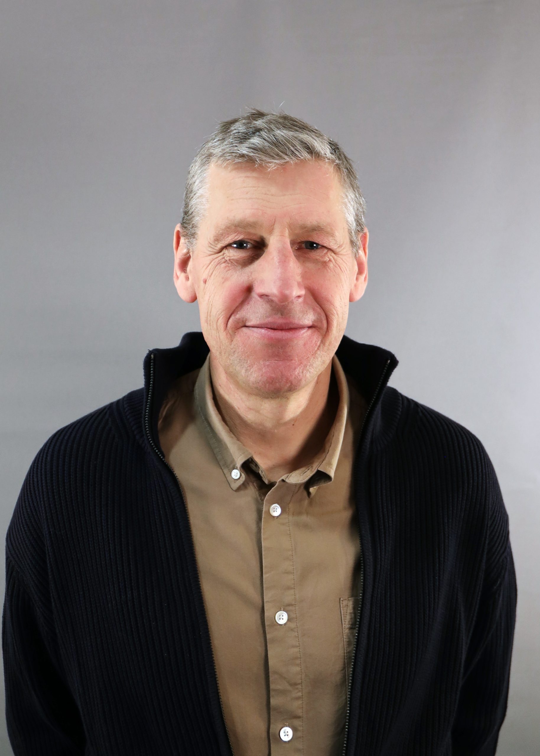 En medelålders man med kort grått hår ler, klädd i brun skjorta och mörk kavaj, mot en grå bakgrund.