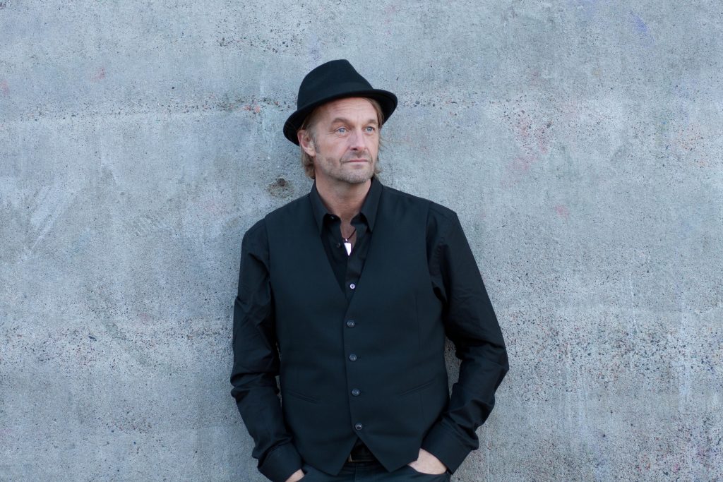 Man i svart skjorta, väst och hatt står mot en grå betongvägg med händerna i fickorna.