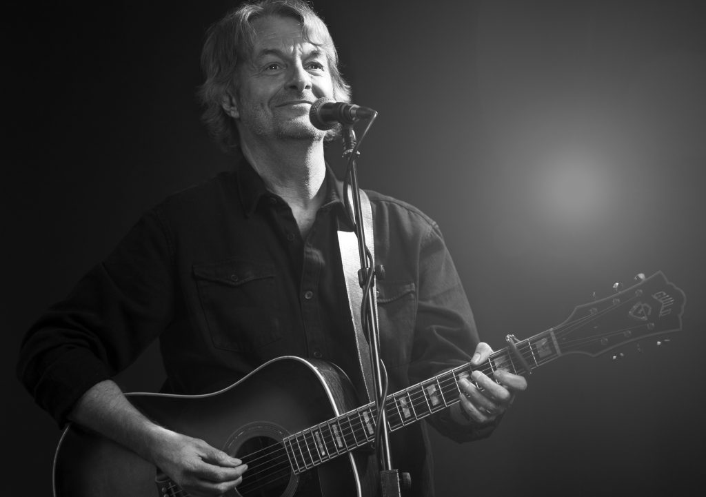 Man spelar akustisk gitarr och sjunger i en mikrofon på scen, svartvitt foto.