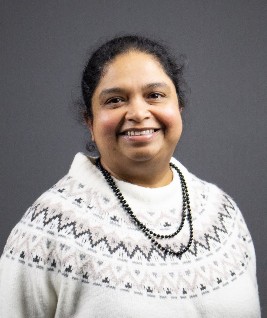 En kvinna i mönstrad vit tröja och svart pärlhalsband ler mot en grå bakgrund.