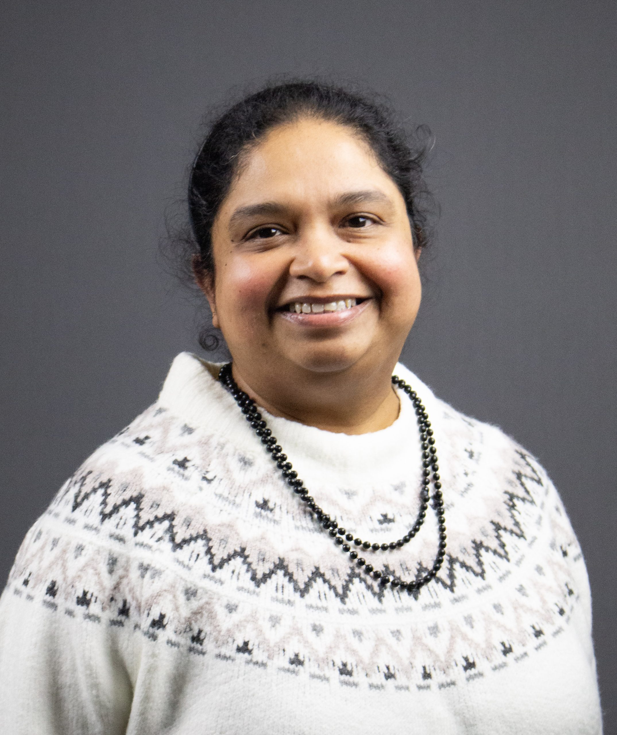 En kvinna i mönstrad vit tröja och svart pärlhalsband ler mot en grå bakgrund.