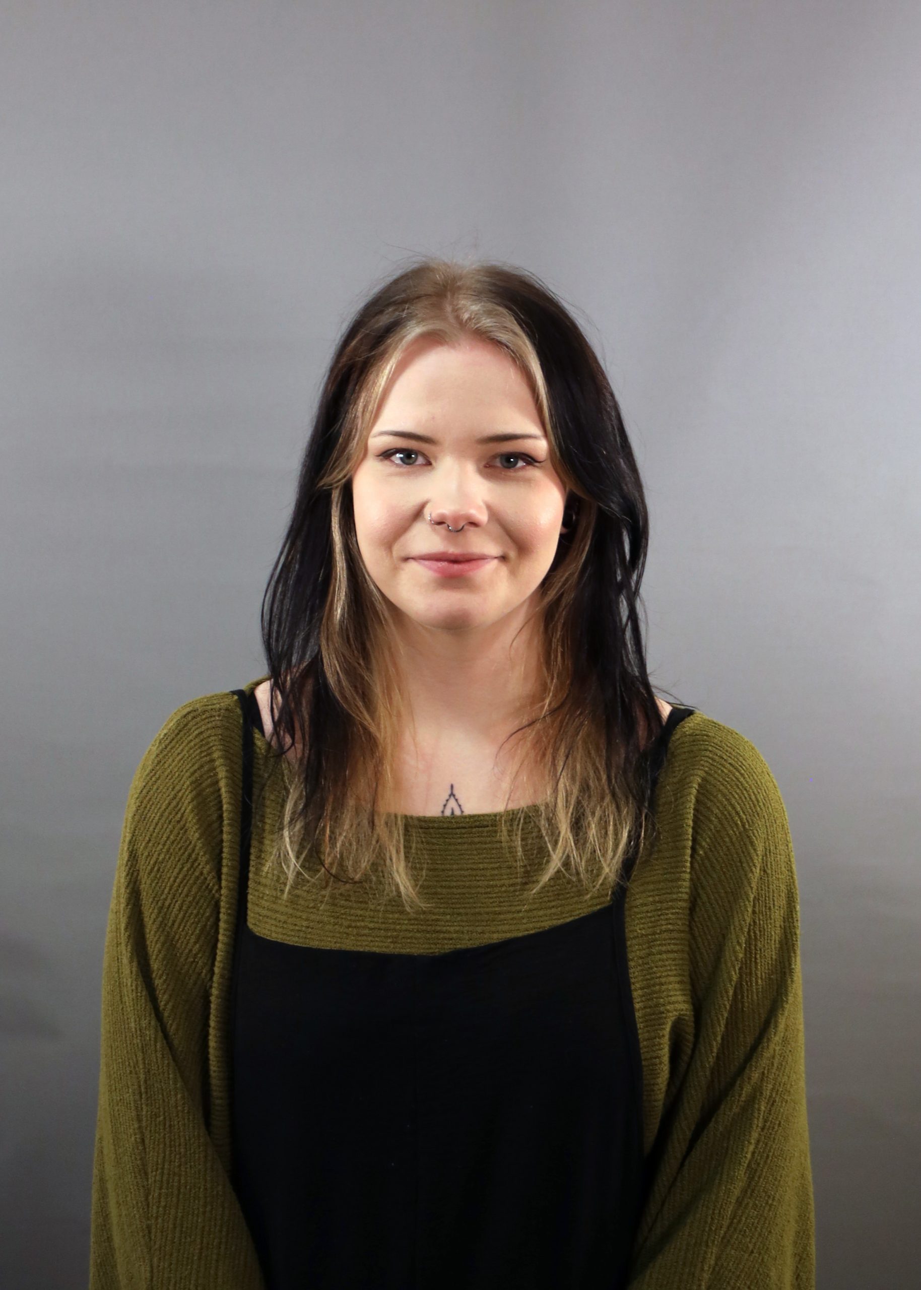 Ung kvinna med långt mörkt hår och grön tröja står framför en grå bakgrund och ler försiktigt.