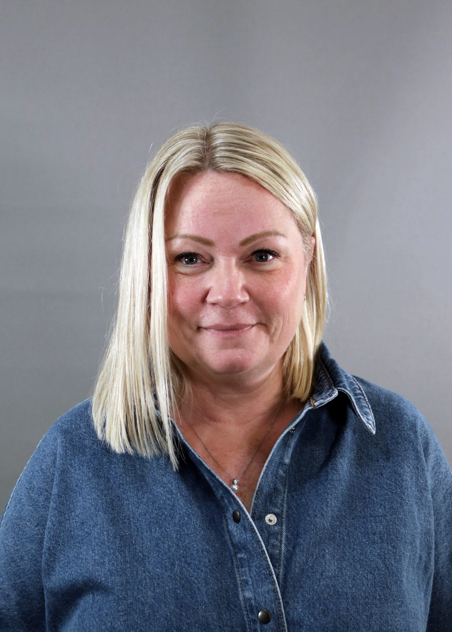 En kvinna med blont hår i jeansskjorta ler mot en grå bakgrund.