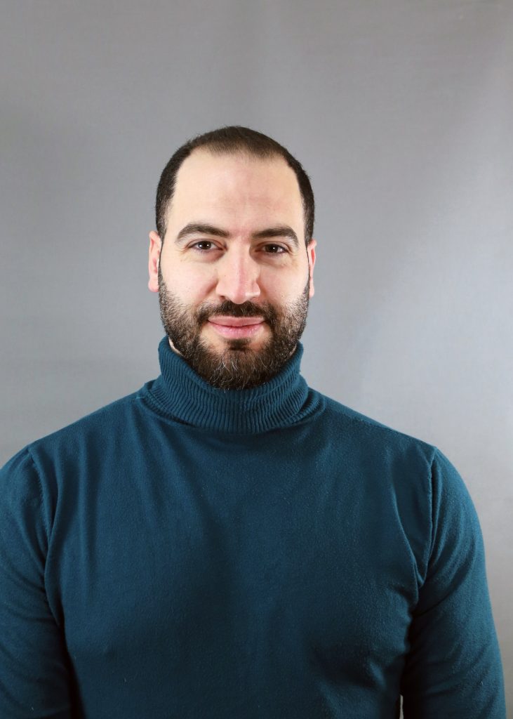 En man med skägg och en blågrön polotröja står framför en grå bakgrund.