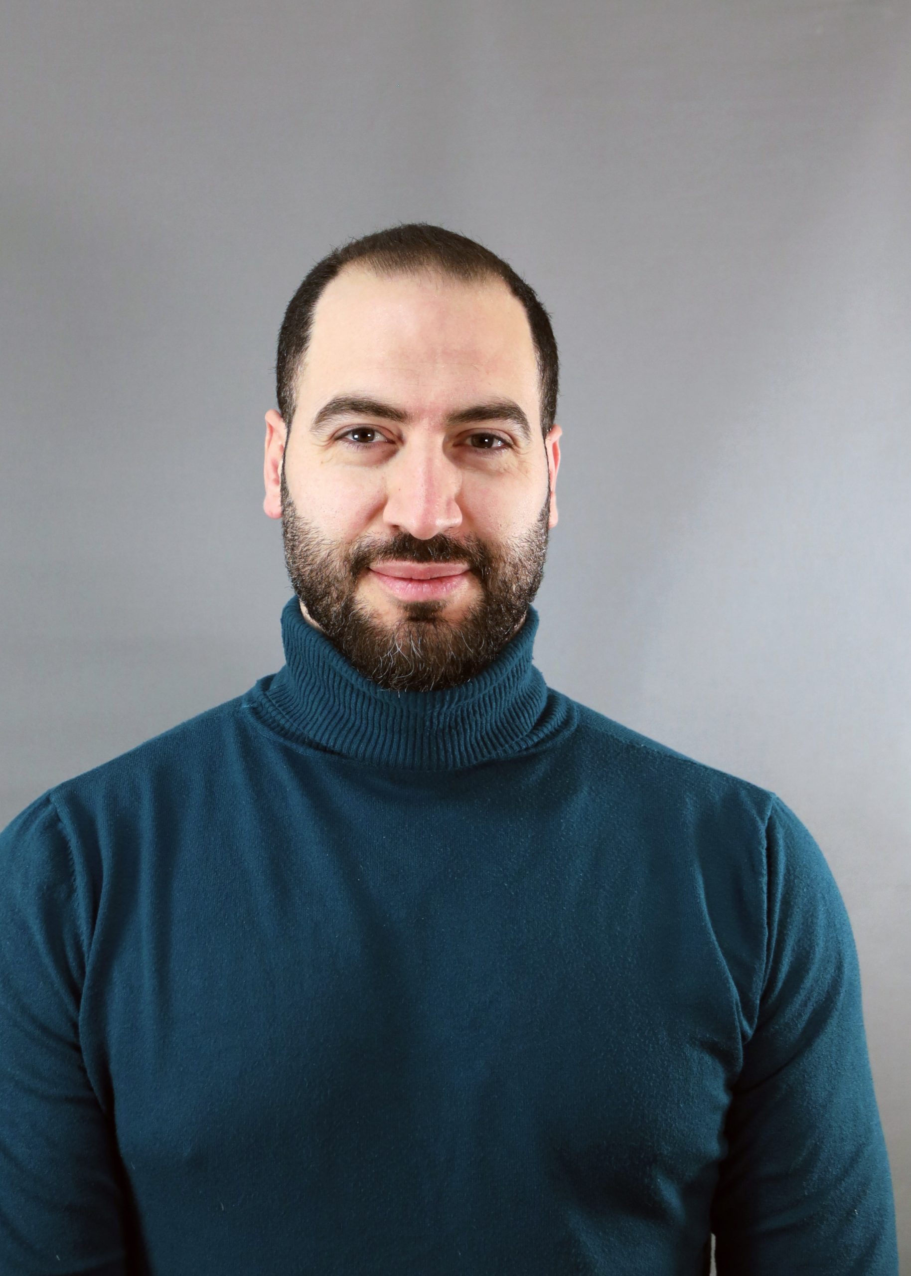 En man med skägg och en blågrön polotröja står framför en grå bakgrund.