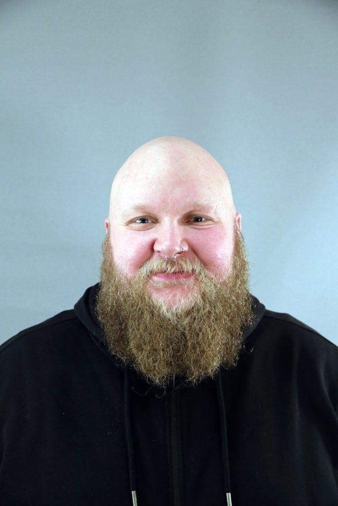 En leende skäggig man med skallig hjässa och svart huvtröja står framför en grå bakgrund.