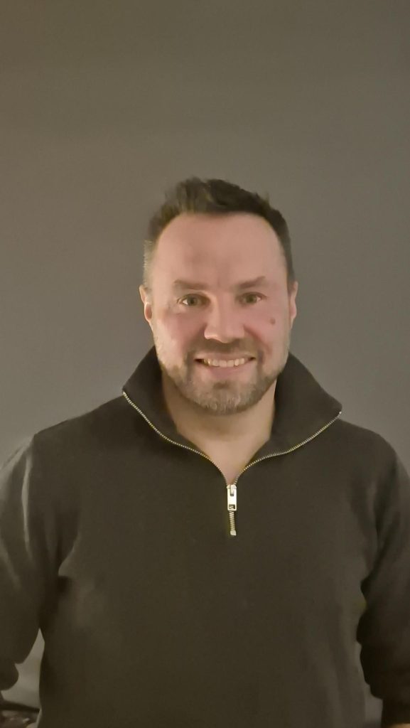 En man med kort hår och skägg ler, iklädd en mörk tröja med dragkedja, mot en enfärgad bakgrund.