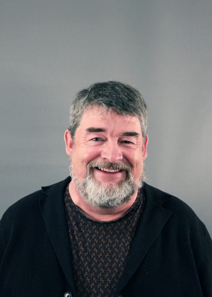 Leende äldre man med grått hår och skägg, iklädd en mörk tröja, mot en grå bakgrund.