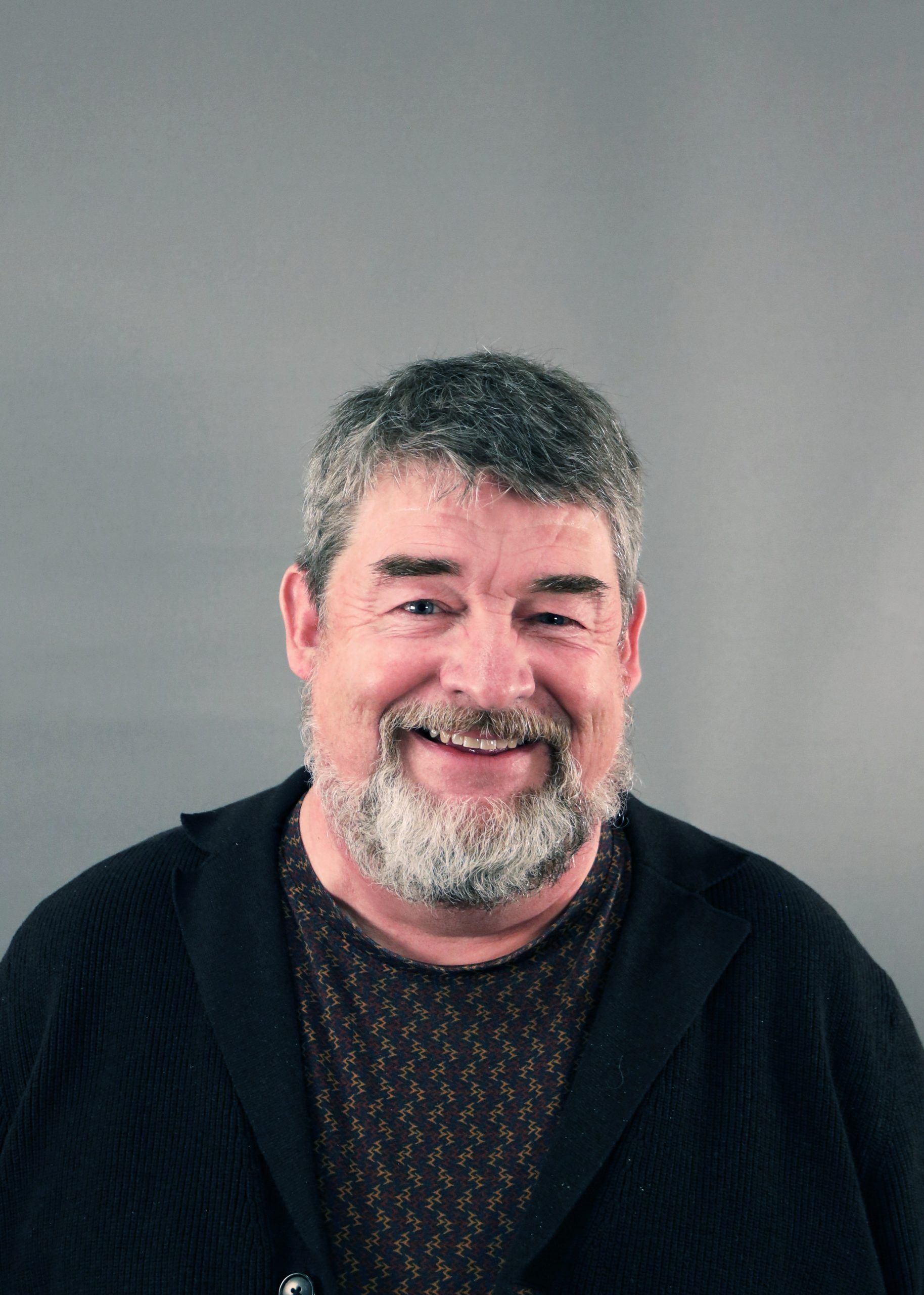 Leende äldre man med grått hår och skägg, iklädd en mörk tröja, mot en grå bakgrund.