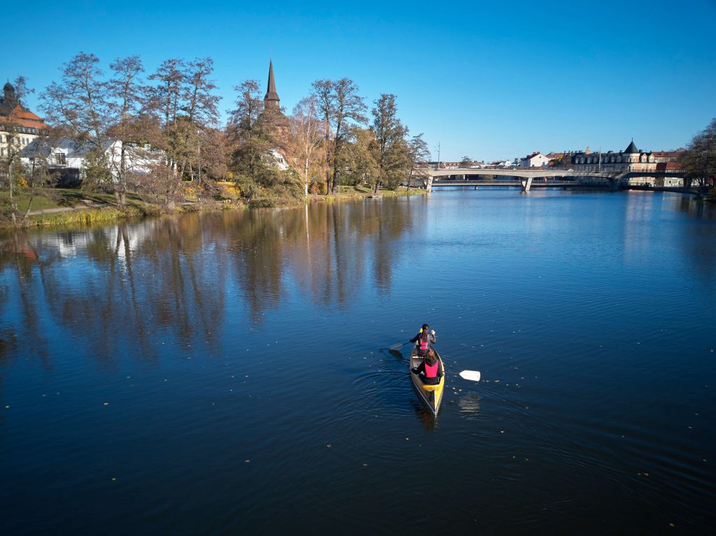 En person paddlar kanot på en lugn flod omgiven av träd och byggnader, med en bro och en kyrkspira i bakgrunden under en klarblå himmel.
