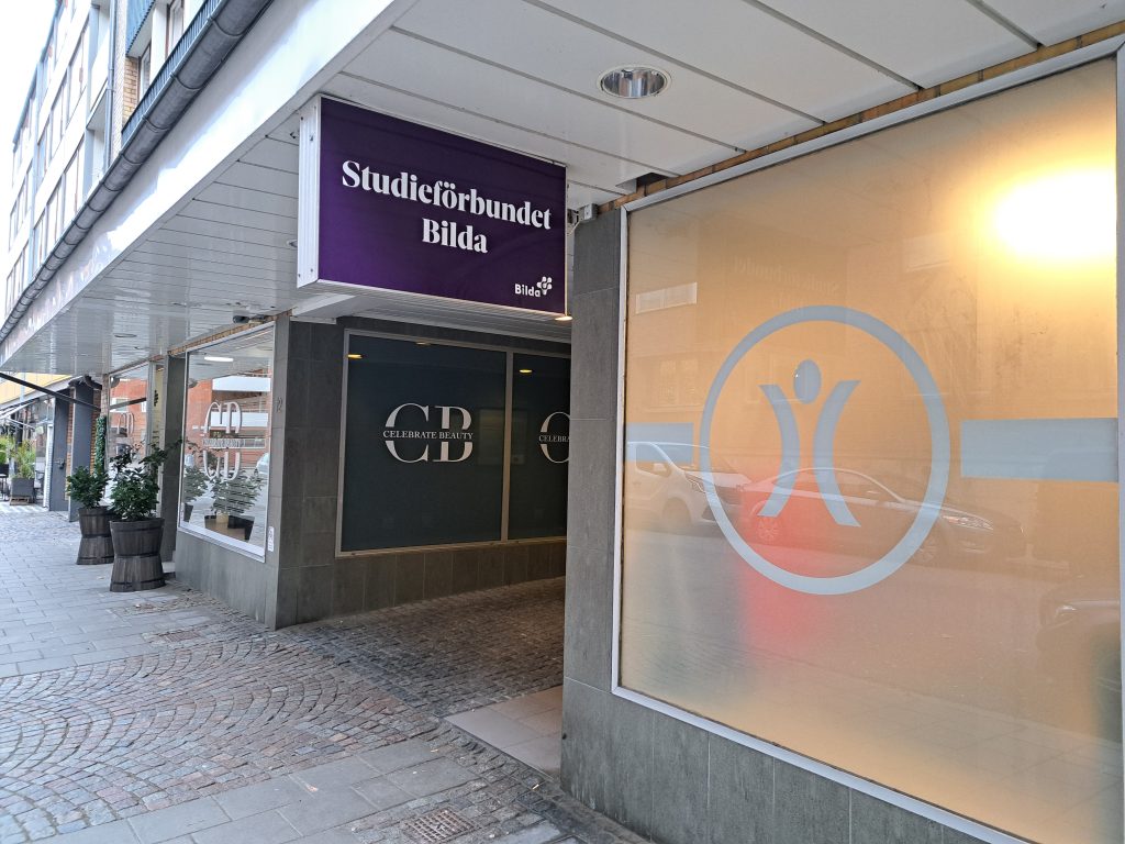 En trottoarvy av en byggnad med en "Studieförbundet Bilda"-skylt och fönster i frostat glas.