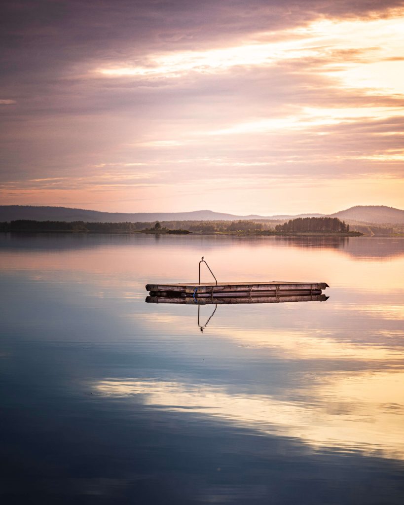 En flytande träbrygga ligger på lugnt, reflekterande vatten under en pastellfärgad himmel vid soluppgång eller solnedgång, med avlägsna kullar och mjuka moln i bakgrunden.