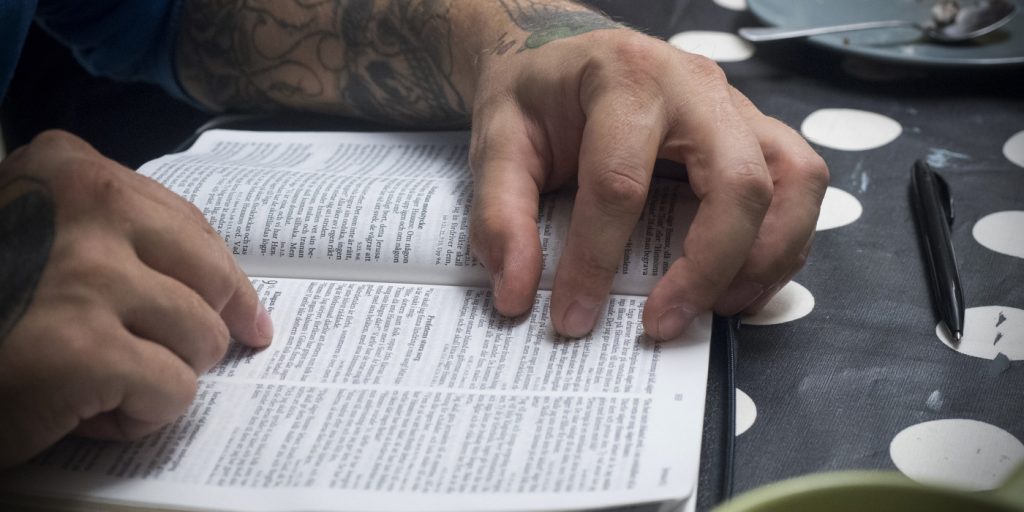 Tatuerade händer läser i en uppslagen bok på ett prickigt bord med penna och disk i närheten.