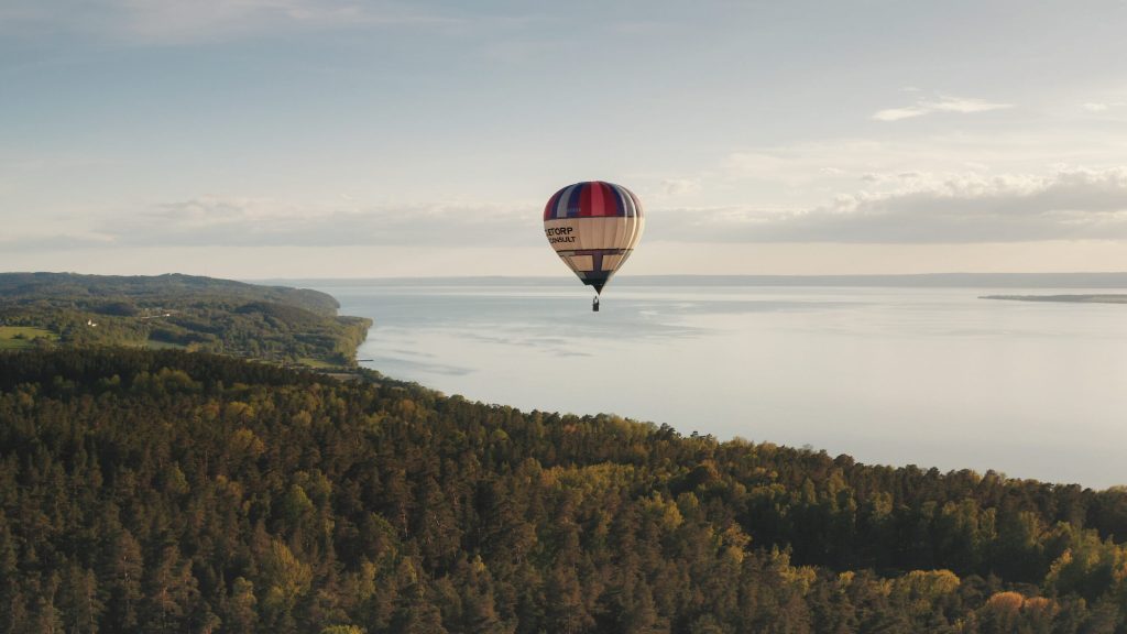 En färgglad varmluftsballong svävar över ett skogslandskap nära en stor vattensamling under en klar himmel med spridda moln.