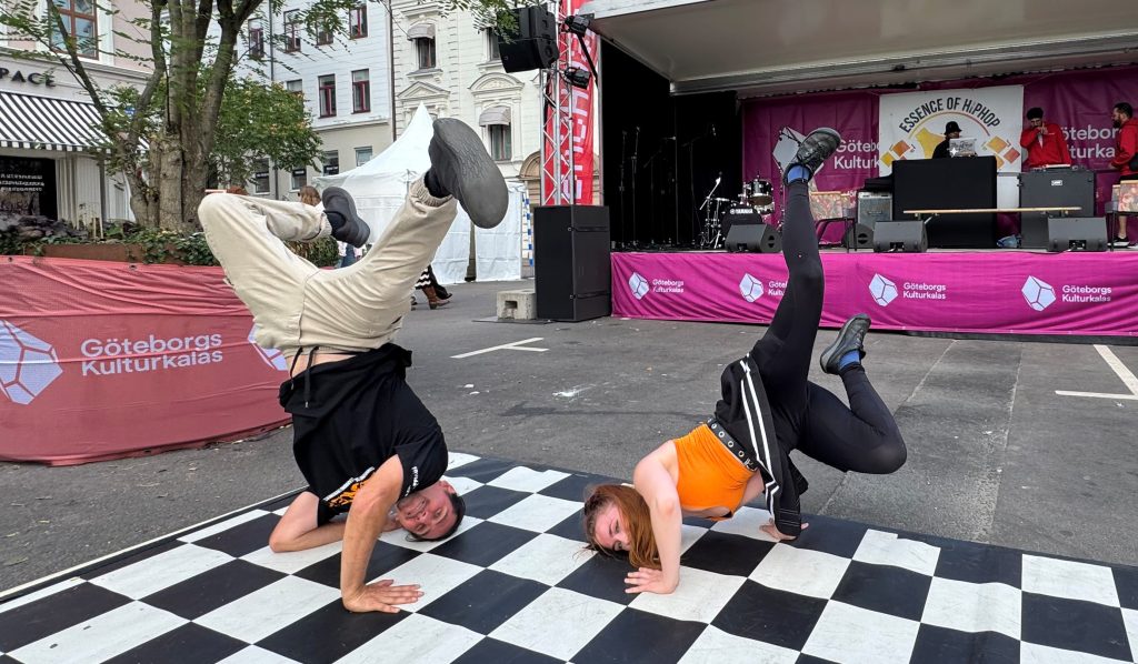 Två personer dansar breakdance på ett rutigt golv på ett utomhusevenemang med en scen i bakgrunden.