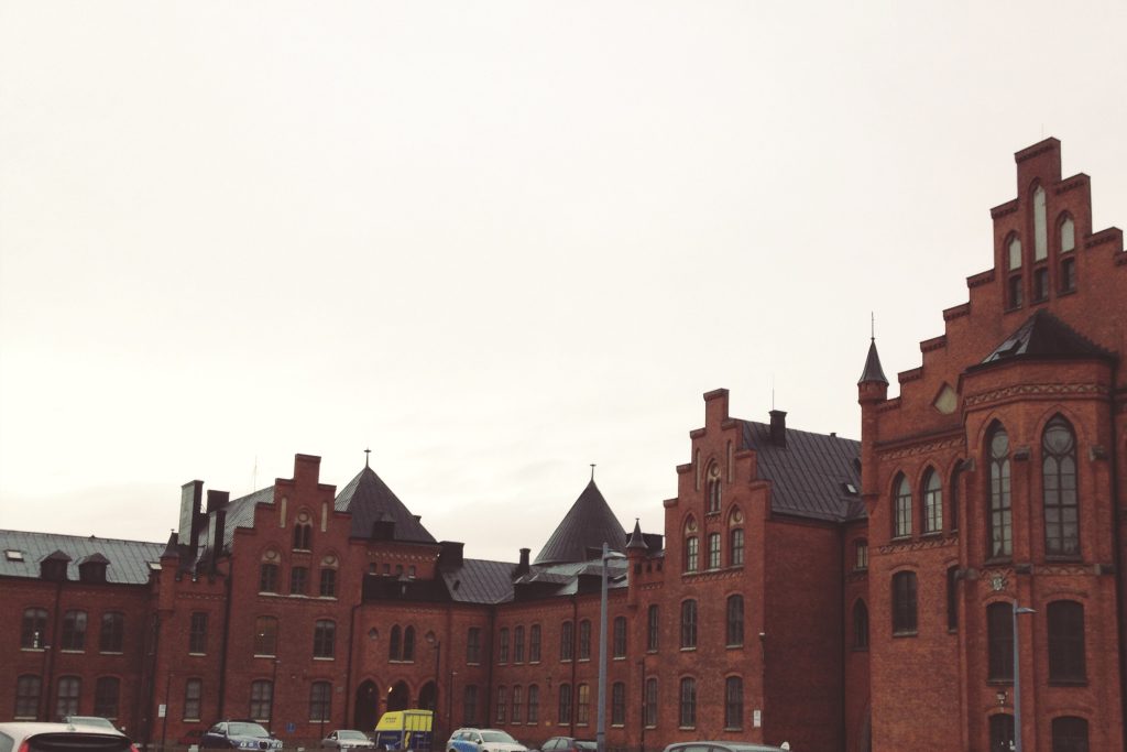 Stor röd tegelbyggnad med sadeltak, välvda fönster och parkerade bilar framför under en molnig himmel.