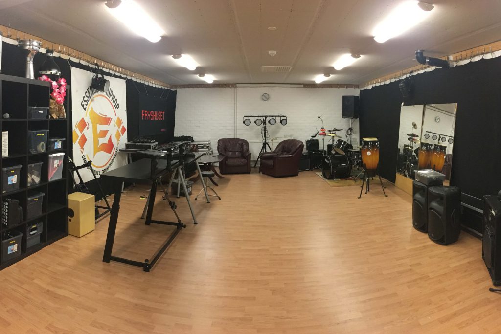 Musikstudio med instrument, stolar, speglar, hyllor och ljudutrustning i ett rymligt rum med trägolv.