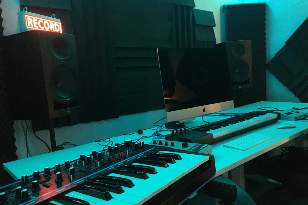En musikstudio med keyboards, högtalare, en iMac och en röd "RECORD"-skylt som lyser upp.