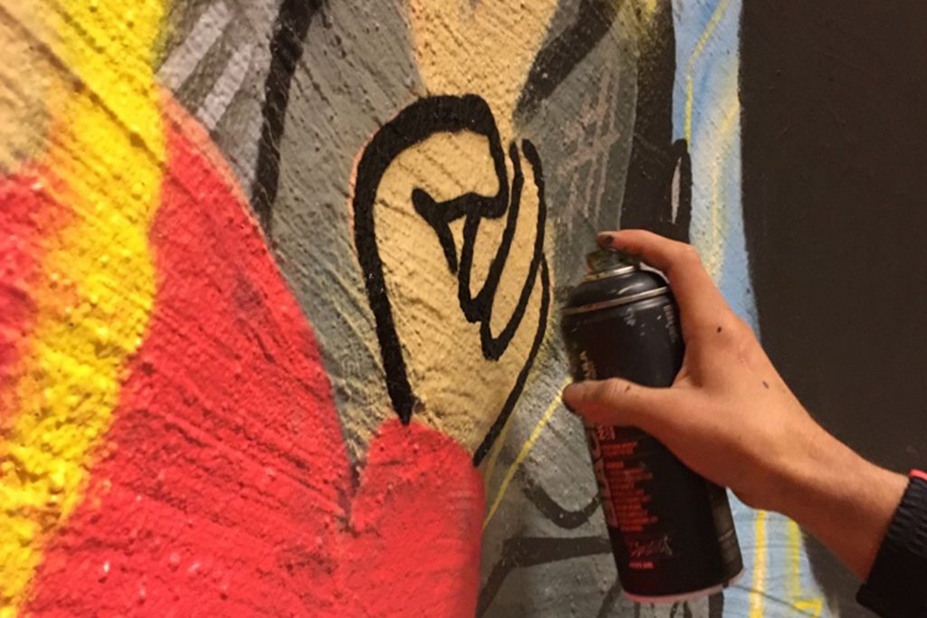 En hand spraymålar svarta linjer över färgglad graffiti på en strukturerad vägg.