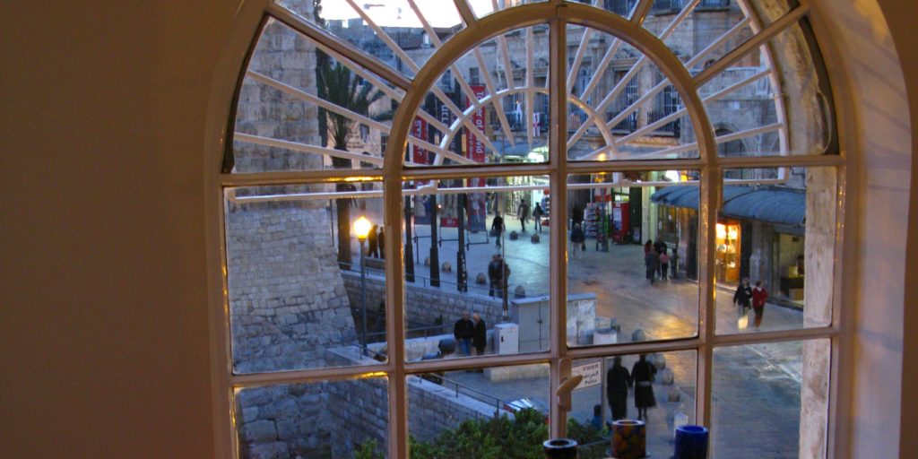 Utsikt över ett stentorg med promenerande människor, sett genom ett stort välvt fönster med radiella rutor.