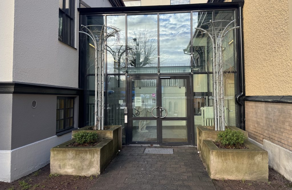 Glasentré med dubbeldörrar, flankerad av två planteringar och bågformade metallkonstruktioner som speglar himlen.