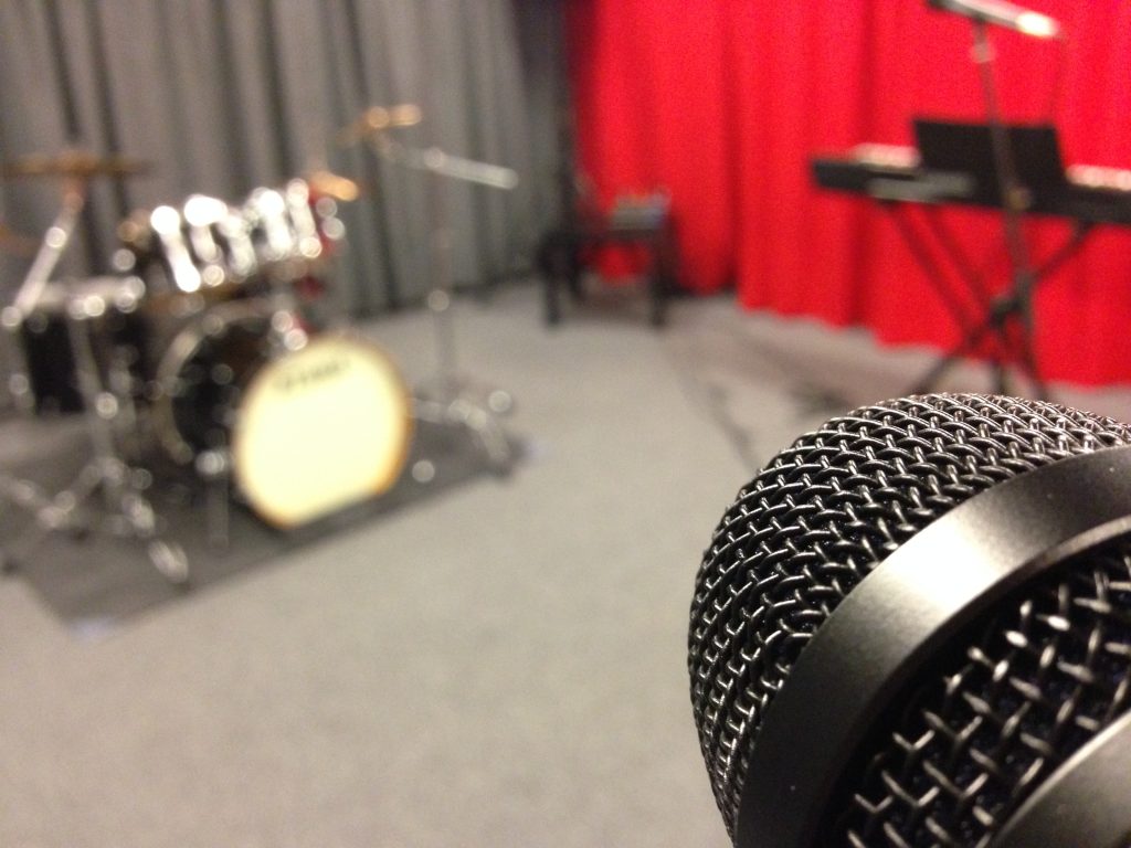 Närbild av en mikrofon med ett suddigt trumset, keyboard och en röd gardin i bakgrunden.