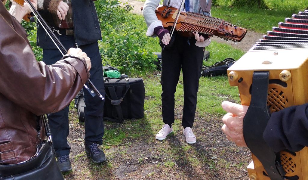 Musiker som spelar sträng- och dragspelsinstrument utomhus, med en bakgrund av gräs och gröna bladverk.