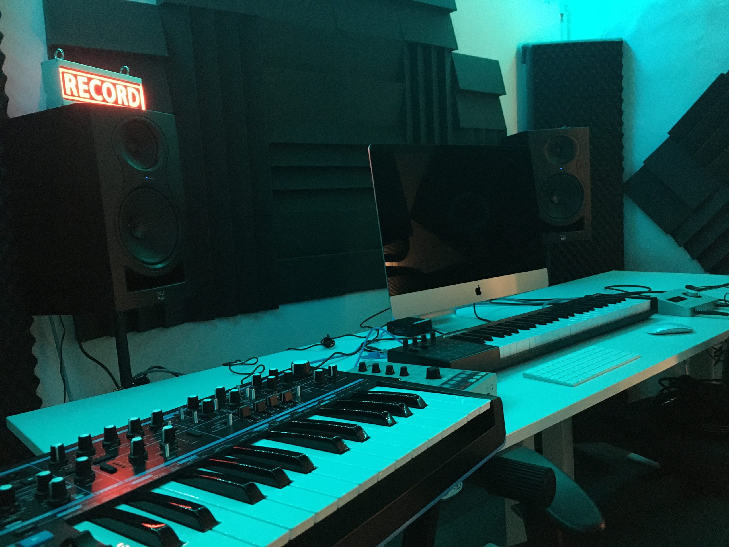 Musikstudio med keyboards, högtalare, dator och en röd skylt med texten "RECORD" på väggen.