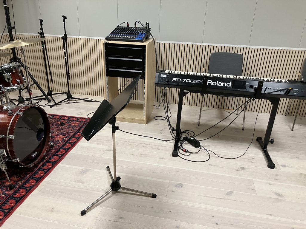 En musikstudio med ett trumset, Roland keyboard, notställ, ljudmixer och kablar på ett ljust trägolv.