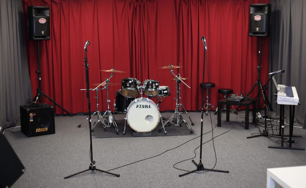 Ett trumset, två mikrofoner, högtalare och en keyboard i ett musikrepetitionsrum med röda gardiner.