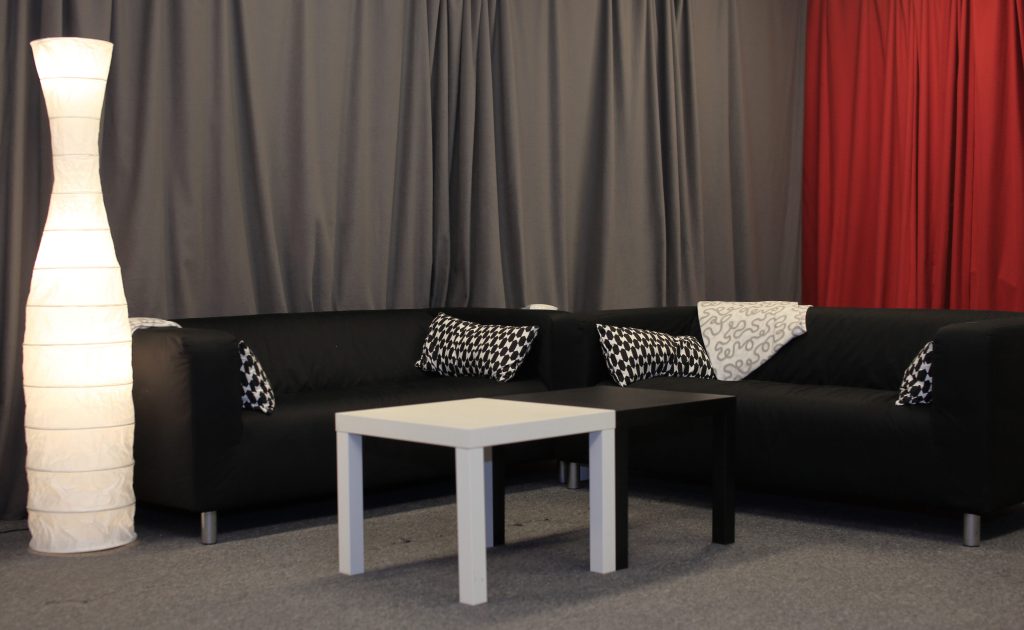 Två svarta soffor, mönstrade kuddar, ett vitt och svart soffbord, en hög lampa samt grå och röda gardiner.