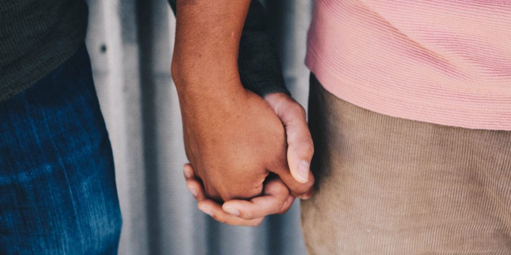 Två personer som håller varandra i handen, den ena klädd i rosa och brunt, den andra i mörka ärmar och blå jeans.