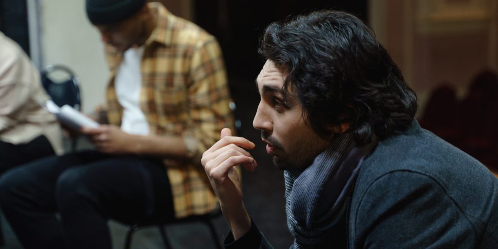 En man med mörkt hår sitter tankfullt medan en annan man i rutig skjorta läser i bakgrunden.