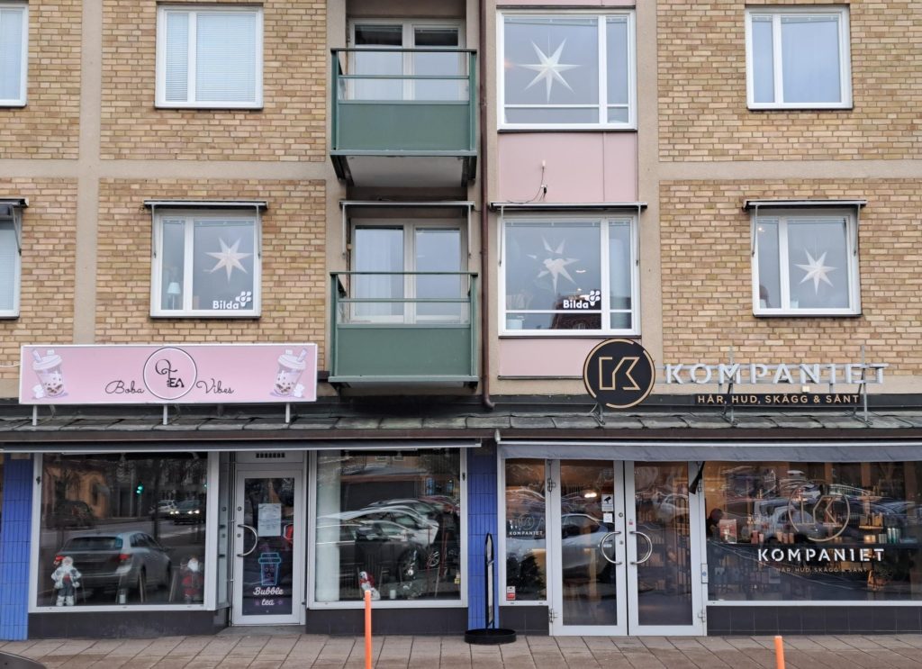 Frontvy av två skyltfönster: Bageriet "Cake n' Vibe" och hår- och hudsalongen "KOMPANIET" i en tegelbyggnad.