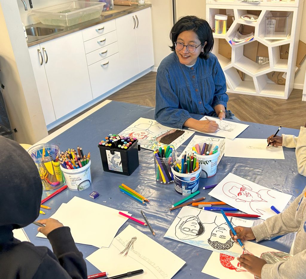 En kvinna sitter vid ett bord med barn och ritar med färgpennor och pennor. Konstmaterial och skisser av ansikten ligger utspridda över bordet.