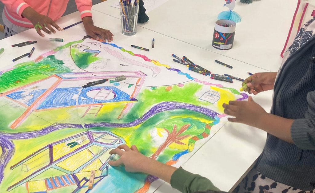Flera barn ritar och färglägger tillsammans ett stort, färgstarkt landskap på vitt papper med hjälp av tuschpennor och kritor på ett bord.