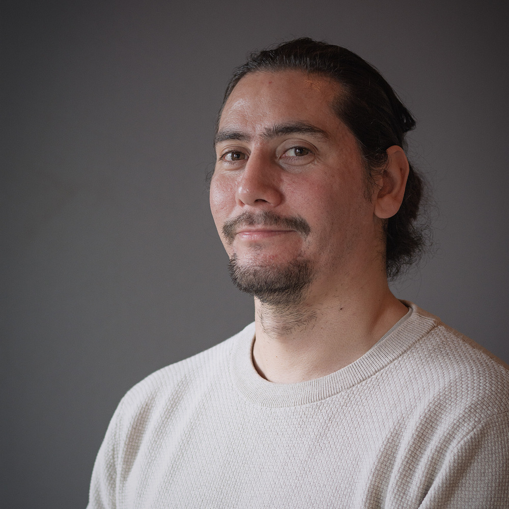 En man med långt mörkt hår och skägg, klädd i en ljus tröja, ler lite mot en grå bakgrund.