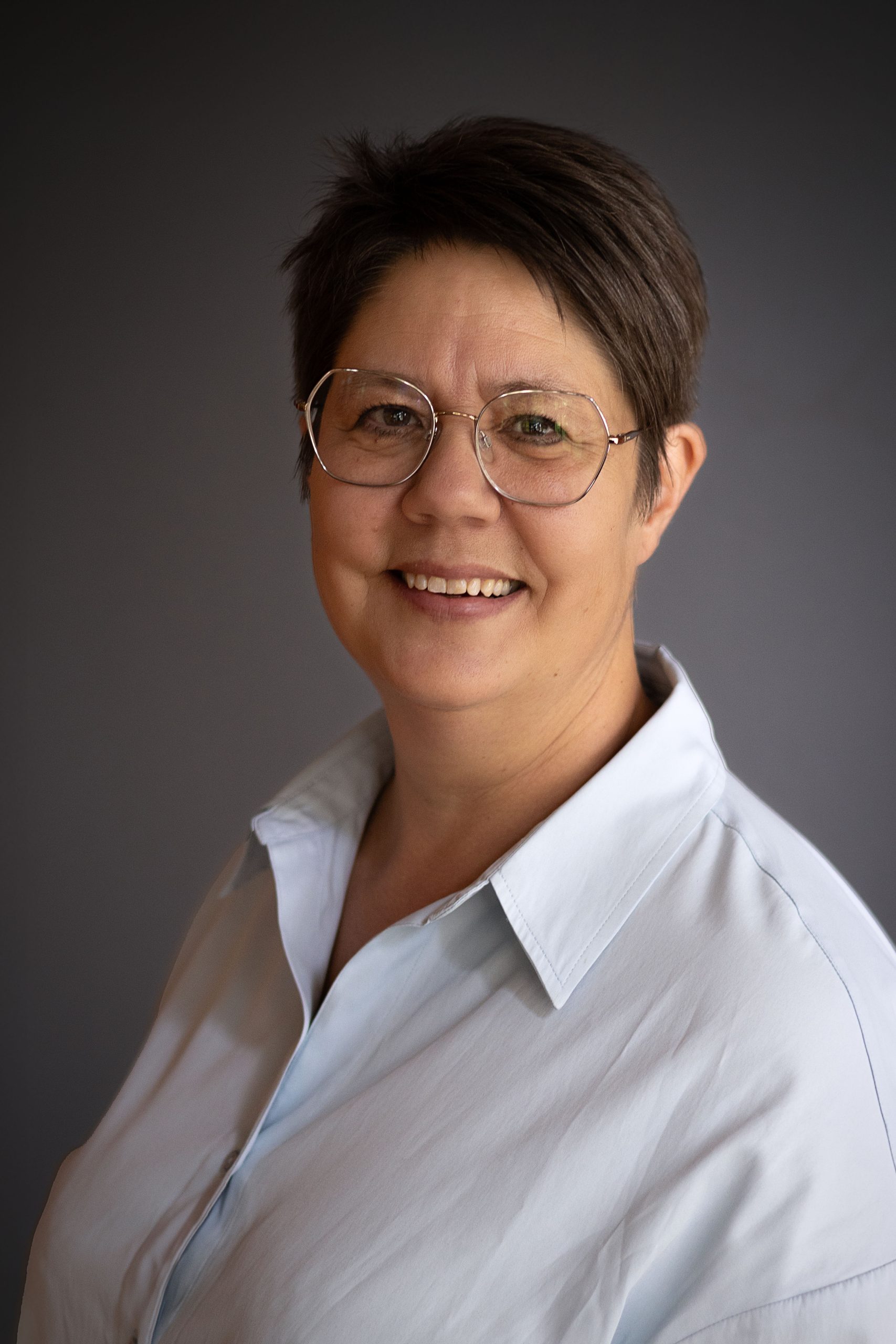 Leende kvinna med kort brunt hår, glasögon och ljusblå skjorta mot en mörk bakgrund.