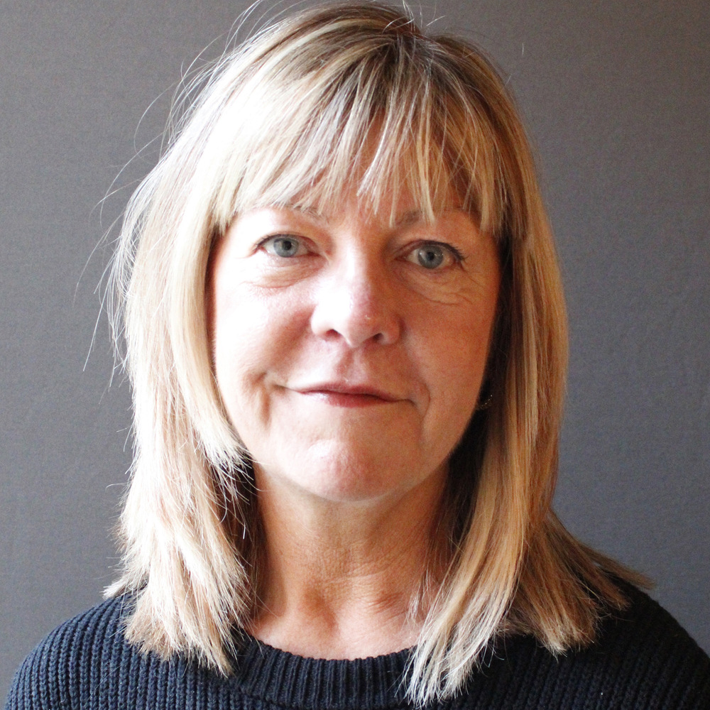 En kvinna med blont hår och lugg, klädd i en svart tröja, ler lätt framför en grå bakgrund.