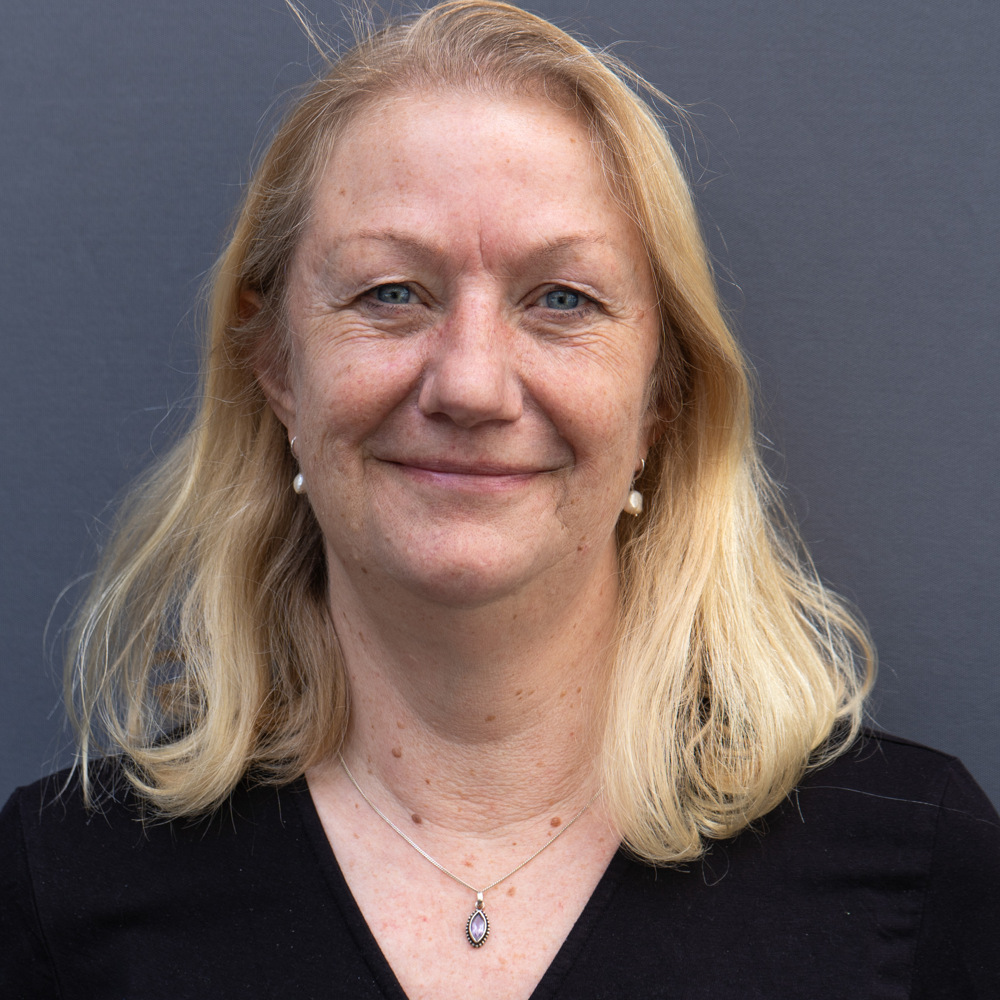 Leende medelålders kvinna med blont hår, klädd i svart topp och halsband, mot grå bakgrund.