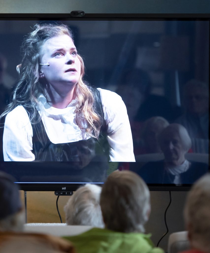 Personer på äldreboende tittar på inspelad teater på stor skärm