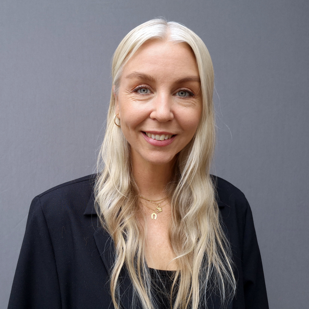 Kvinna med långt blont hår, svart kavaj och ett leende framför en grå bakgrund.